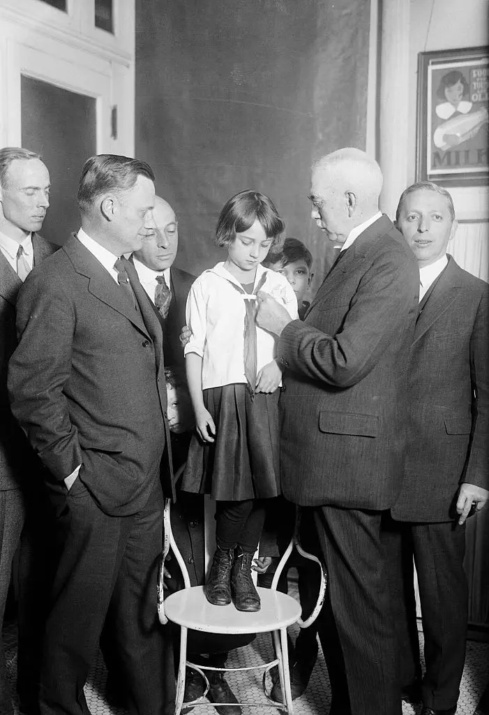   فتاة وهي تتلقى التطعيم ضد الدفتيريا. في الصورة، يقوم الدكتور فرانك ج. موناغان، مفوض الصحة بمدينة نيويورك، بوضع شارة التطعيم على قميص الفتاة 